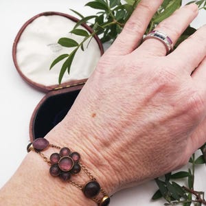 May include: A vintage gold bracelet featuring amethyst-coloured stones and a floral design. The bracelet is worn on a wrist, with a ring on a finger. A jewelry box is in the background.