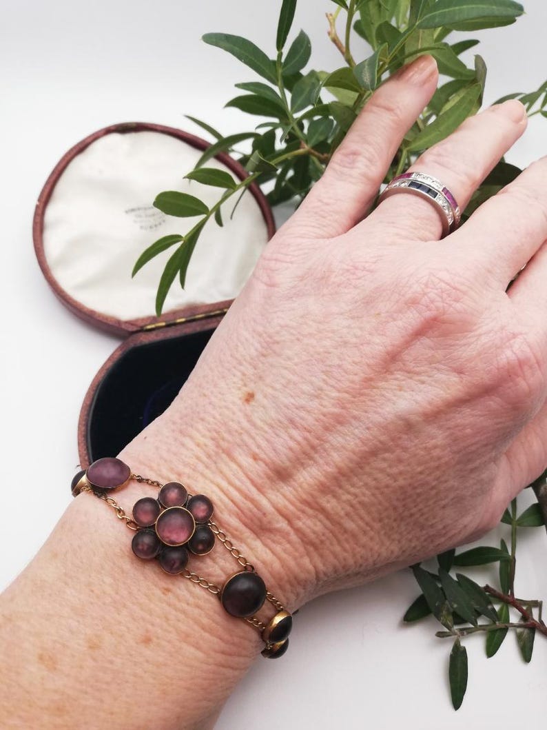 May include: A vintage gold bracelet featuring amethyst-coloured stones and a floral design. The bracelet is worn on a wrist, with a ring on a finger. A jewelry box is in the background.
