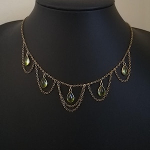 May include: A gold-coloured necklace featuring multiple strands and teardrop-shaped green gemstones. The necklace is displayed on a black mannequin, highlighting its delicate design and intricate chainwork. The gemstones are evenly spaced along the necklace.