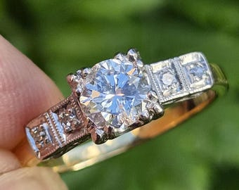 Pode incluir: Um anel de ouro com uma grande pedra central redonda de corte brilhante num engaste de metal branco. A banda inclui duas pedras de destaque mais pequenas de cada lado, inseridas em secções retangulares com detalhes de milgrain.