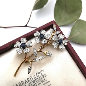 May include: A gold and silver floral brooch with three blue sapphires and diamonds. The brooch is in a red velvet box with the text "GARRARD & CO. LTD CROWN JEWELLERS REGENT ST.W.I."