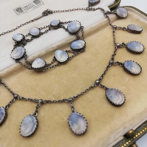 May include: A silver necklace and bracelet set featuring oval-shaped, pale blue, cameo-like stones. The necklace has a chain with five dangling stones and the bracelet has a double chain with six stones. The set is presented in a green velvet lined box with a label that reads "Liberty & Co. London".