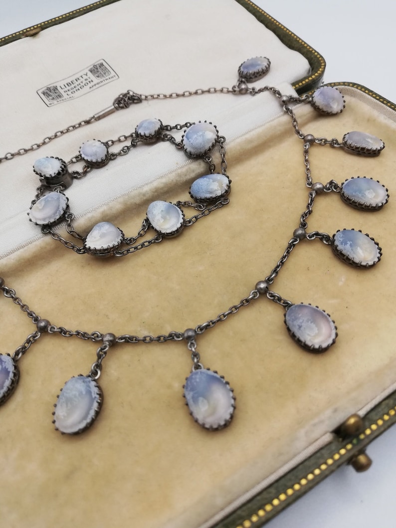May include: A silver necklace and bracelet set featuring oval-shaped, pale blue, cameo-like stones. The necklace has a chain with five dangling stones and the bracelet has a double chain with six stones. The set is presented in a green velvet lined box with a label that reads "Liberty & Co. London".