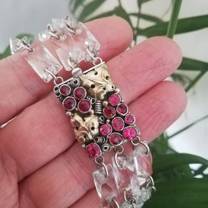 Puede incluir: Un brazalete plateado y dorado con cristales rectangulares transparentes y piedras preciosas rojas. El brazalete presenta una sección central dorada con detalles intrincados, conectada por eslabones plateados.