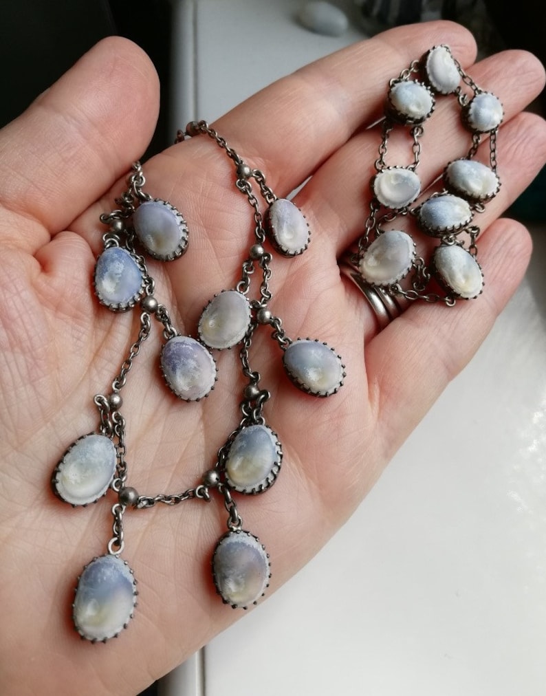May include: A silver chain necklace with multiple strands and oval-shaped pendants. The pendants are made of a white, iridescent material with a silver trim.