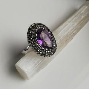 May include: Silver ring featuring an oval amethyst gemstone, framed by marcasite stones. The ring's design is set on a white, textured surface. The amethyst is a vibrant purple colour, and the ring is a statement piece.