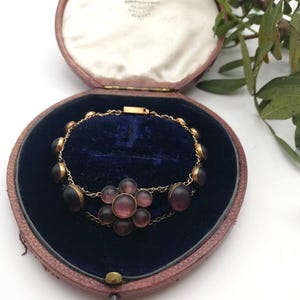 May include: A vintage gold bracelet featuring dark purple oval stones and a flower-shaped cluster of purple stones. The bracelet is presented in an open, heart-shaped, red leather jewellery box with a blue velvet interior.