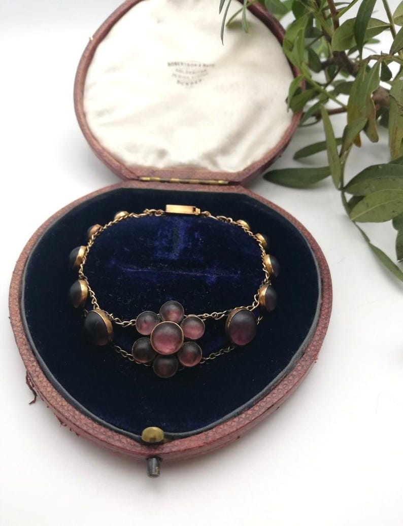 May include: A vintage gold bracelet featuring dark purple oval stones and a flower-shaped cluster of purple stones. The bracelet is presented in an open, heart-shaped, red leather jewellery box with a blue velvet interior.