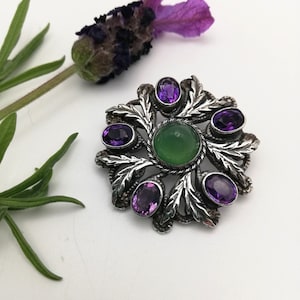 May include: A silver brooch featuring a central green stone surrounded by six purple stones. The brooch is designed in the shape of a flower with leaves.