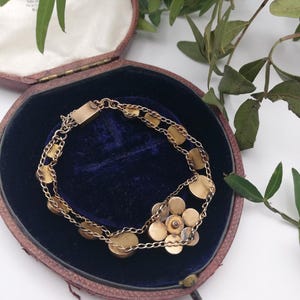 May include: A gold bracelet featuring a floral cluster and circular links, presented in a vintage maroon jewellery box with a blue velvet lining. Green foliage adds detail.