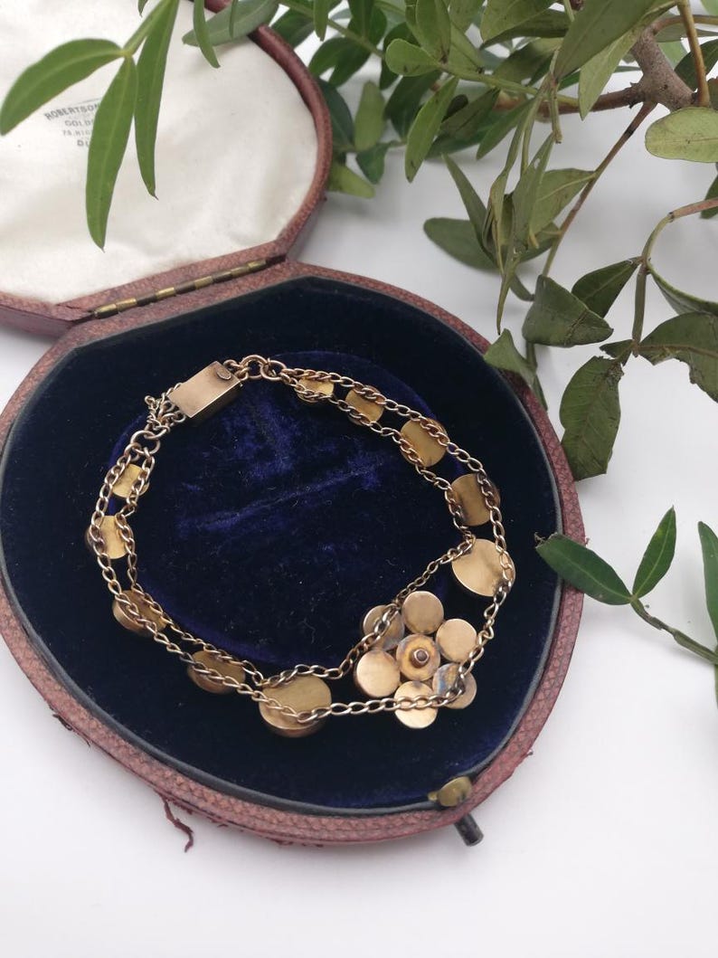May include: A gold bracelet featuring a floral cluster and circular links, presented in a vintage maroon jewellery box with a blue velvet lining. Green foliage adds detail.