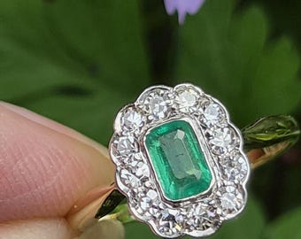 May include: A ring featuring a central emerald-cut green gemstone surrounded by a scalloped halo of small round white diamonds. The setting is crafted from a metallic band, showcasing a classic cluster design with a vibrant green center stone.