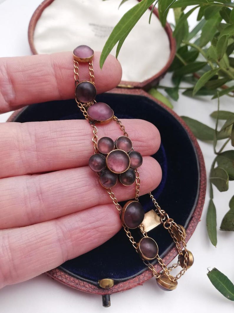 May include: A vintage gold bracelet with mauve-coloured oval and circular stones, arranged in a floral pattern. The bracelet is displayed in a vintage jewellery box with a blue interior and a pink exterior.