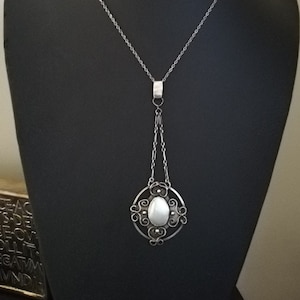 May include: A silver necklace with a delicate chain and a pendant featuring a white oval stone surrounded by a silver filigree design.