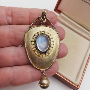 May include: A teardrop-shaped pendant with a brushed gold-tone finish featuring a central oval cabochon stone with a milky blue sheen. The pendant is framed by a decorative scalloped border and includes a small spherical drop charm at the base.