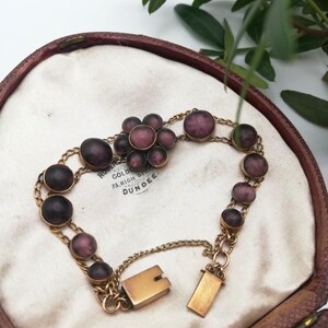 May include: A vintage gold bracelet featuring purple, oval and round stones. The bracelet is presented in an open, brown jewellery box with a white interior. The clasp is open, and the chain is delicate.