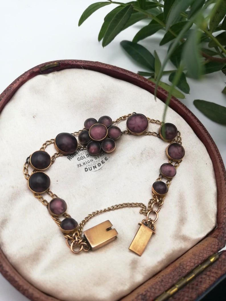 May include: A vintage gold bracelet featuring purple, oval and round stones. The bracelet is presented in an open, brown jewellery box with a white interior. The clasp is open, and the chain is delicate.