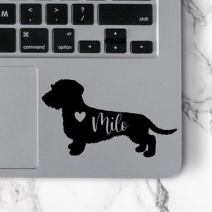 May include: Black silhouette of a dachshund dog with a heart and the name "Milo" on a silver laptop.