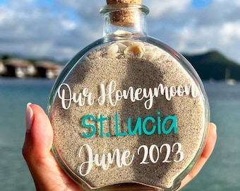 May include: A round glass bottle with a cork stopper, filled with light beige sand and small white seashells. The front of the bottle features white and teal text that reads: Our Honeymoon, St. Lucia, June 2023, printed in a script and sans-serif font.