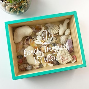 May include: A teal wooden box with a glass top filled with seashells and coral. The box has the text "Beach Treasures Key West, FL" written on it in white.