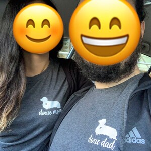 May include: A couple wearing matching gray t-shirts with a dachshund silhouette and the text "doxie mom" and "doxie dad". The man is wearing a black Adidas jacket.
