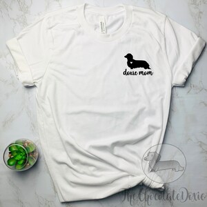 May include: White t-shirt with a black silhouette of a dachshund with a heart on its chest and the text "doxie mom" printed on the front.