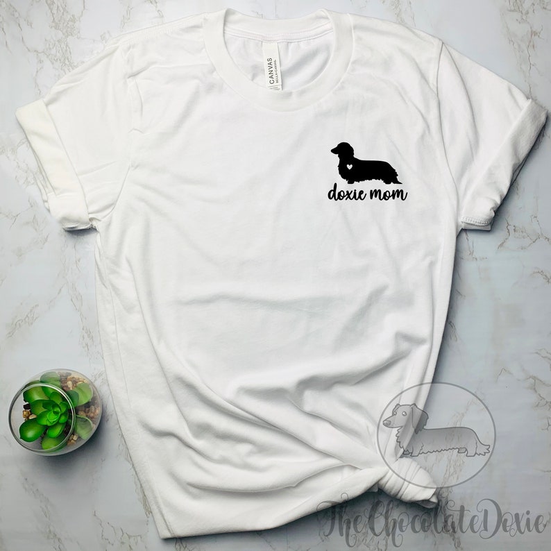 May include: White t-shirt with a black silhouette of a dachshund with a heart on its chest and the text "doxie mom" printed on the front.
