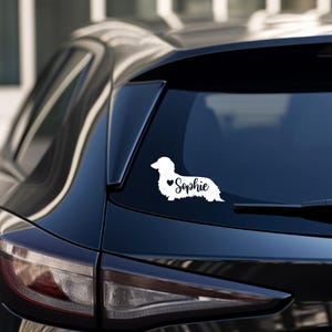 May include: White vinyl decal of a dachshund silhouette with a small heart and the name "Sophie" on a black car.