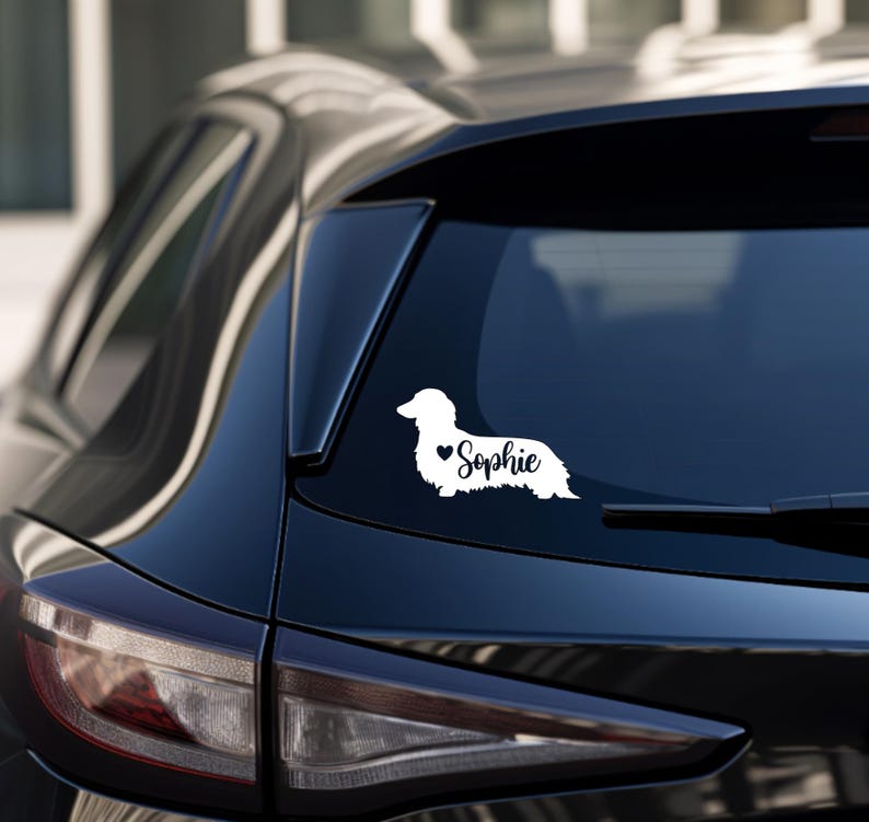 May include: White vinyl decal of a dachshund silhouette with a small heart and the name "Sophie" on a black car.