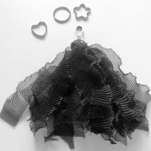 May include: A black, pleated fabric accessory with a silver clasp, alongside three small, silver-coloured metal shapes: a heart, a circle, and a star. The fabric has a layered, ruffled appearance.