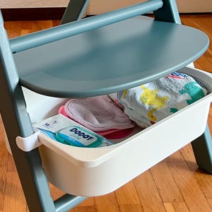 Könnte beinhalten: Hellblauer und grauer Hochstuhl mit einer weißen Aufbewahrungsbox. Die Box enthält Babytücher, ein rosa Tuch und ein gefaltetes Handtuch mit gelben und grünen Designs. Der Stuhl steht auf einem Parkettboden.
