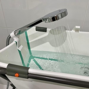 Może przedstawiać: Biała i zielona przenośna wanienka dla dziecka z chromowaną słuchawką prysznicową rozpylającą wodę. Wanienka ma szarą ramę i pomarańczowy przycisk. Woda jest czysta, a tło to biała kafelkowa ściana.