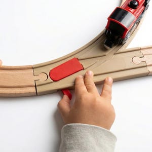 Pode incluir: Um comboio de brinquedo vermelho sobre uma linha de madeira com uma mudança de agulha. A linha é castanha clara com uma secção de mudança de agulha bege. As mãos de uma criança ajustam a mudança. O comboio é vermelho e preto.