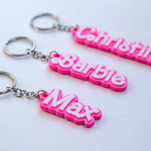 Puede incluir: Tres llaveros rosas y blancos con nombres. Los nombres "Christina", "Barbie" y "Max" se muestran en letras blancas en relieve sobre los llaveros rosas. Cada llavero tiene un anillo plateado.