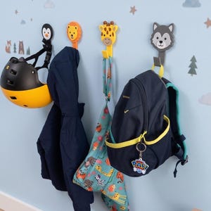 Peut inclure: Quatre patères murales en forme de têtes d'animaux, représentant un pingouin, un lion, une girafe et un loup. Fixées sur un mur bleu clair, elles supportent un casque jaune et noir, une veste bleu marine, un sac à motifs et un sac à dos.