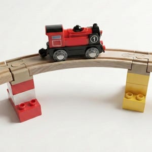 Könnte beinhalten: Ein roter Spielzeugzug mit der Nummer 1 an der Seite befindet sich auf einer Holzbrücke, die von roten, weißen und gelben Spielzeugblöcken getragen wird. Der Zug ist rot und schwarz mit silbernen Rädern. Die Strecke ist gebogen.