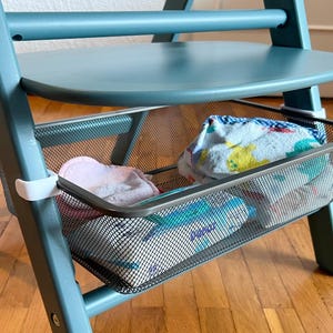 Puede incluir: Una trona de color turquesa con un asiento redondo y una cesta de almacenamiento de malla. La cesta contiene artículos para bebés doblados, incluyendo un paño rosa y una toalla estampada. La silla está sobre un suelo de madera.