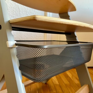 Könnte beinhalten: Ein grauer Holz-Hochstuhl mit einem grauen, geflochtenen Metall-Ablagekorb, der an der Unterseite befestigt ist. Der Stuhl hat eine obere Ablage aus Holz.