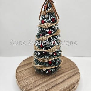 May include: A small, flocked Christmas tree decorated with red berries, white beads, and twine. The tree is on a wooden base with a burlap trim.
