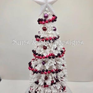 May include: A white flocked artificial Christmas tree decorated with pink and purple beaded garland. The tree is topped with a white glitter star.
