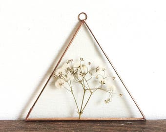 Op de afbeelding: Een driehoekig glas- en koperen frame met een takje gedroogde witte bloemen. Het frame wordt opgehangen door een kleine koperen ring aan de bovenkant. De achtergrond is een witte muur en een houten oppervlak.