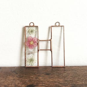 以下が含まれることがあります： 銅線で囲まれたHの文字の中に、押し花が入っています。花は白とピンクです。