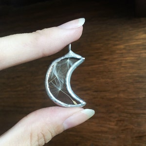 May include: A silver crescent moon pendant with a delicate dandelion inside. The pendant is held by a finger.