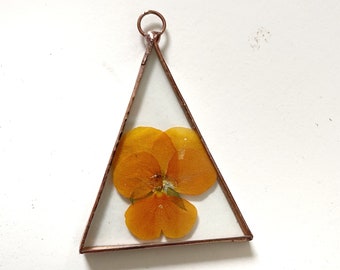 Op de afbeelding: Een koperen driehoekig frame met een geperste oranje bloem erin. Het frame heeft een lus aan de bovenkant om op te hangen.