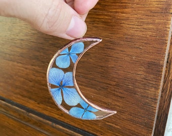 May include: A crescent moon-shaped resin ornament with a copper wire frame. The ornament is filled with pressed blue flowers.