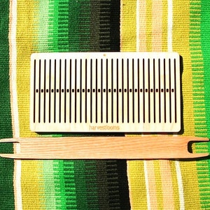 May include: A wooden weaving loom and shuttle. The loom is rectangular with vertical slots and the text "harvestlooms". The shuttle is a long, flat piece of wood with a forked end. The set is on a green, yellow, and white striped textile.