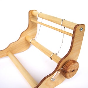K&ouml;nnte beinhalten: Ein Webstuhl aus Holz mit zwei Holzspulen und einem Spannknopf. Der Webstuhl ist aus hellem Holz gefertigt und hat eine nat&uuml;rliche Oberfl&auml;che.