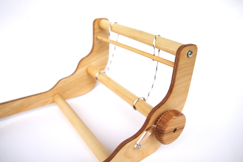 K&ouml;nnte beinhalten: Ein Webstuhl aus Holz mit zwei Holzspulen und einem Spannknopf. Der Webstuhl ist aus hellem Holz gefertigt und hat eine nat&uuml;rliche Oberfl&auml;che.