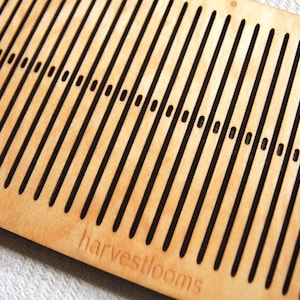 May include: A wooden loom with a series of parallel slots for weaving. The loom is made of light brown wood and has a black inscription that reads "harvestlooms".