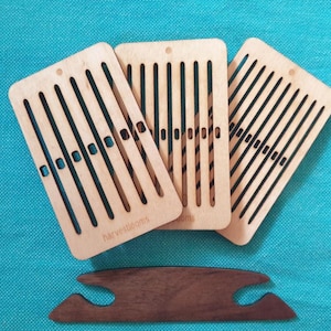 May include: Three light-colored wooden weaving looms with black lines and a dark brown shuttle are displayed on a teal background. The looms have a rectangular shape with a series of vertical slots. The word "harvestlooms" is printed on one of the looms.